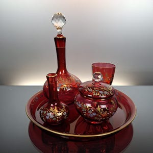 Pode incluir: Um conjunto de vidro vermelho rubi composto por um decantador com rolha de cristal facetado, um vaso pequeno, um frasco com tampa e puxador de cristal e um copo a condizer, dispostos num tabuleiro circular. Cada peça é decorada com esmalte floral e ouro.