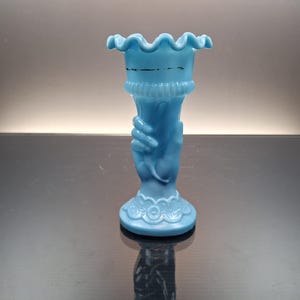 May include: A light blue milk glass vase featuring a sculpted hand design forming the stem. The vase has a ruffled, flared rim and a decorative embossed base. A thin black horizontal line is visible on the upper portion of the glass body.