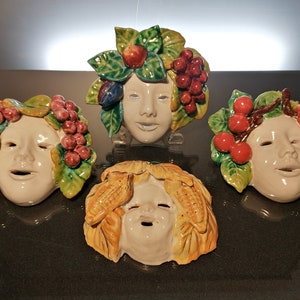Peut inclure: Quatre masques en céramique avec des visages et des décorations de fruits et de légumes. Les masques sont blancs avec des détails peints. Un masque a des raisins, un autre a des cerises, un autre a des pommes et des prunes, et le dernier a du maïs.