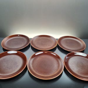 Może przedstawiać: Zestaw sześciu okrągłych, ciemnobrązowych talerzy ceramicznych ułożonych w dwa rzędy po trzy sztuki. Każdy talerz ma gładkie, błyszczące wykończenie z nieco ciemniejszym brzegiem i jaśniejszym, ciepłym brązowym środkiem.