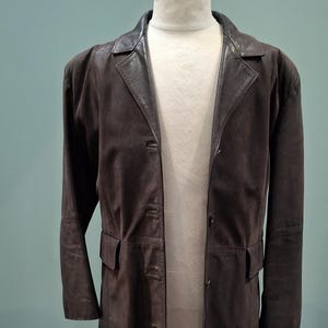 May include: A long brown leather coat with a belt and button fastening. The coat has a classic design and is made from high-quality leather.