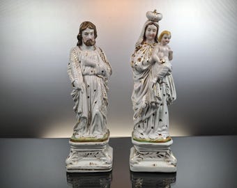 May include: Two white porcelain religious statues. One depicts a man with a beard, the other a woman holding a child. Both figures are adorned with gold accents and stand on ornate bases.