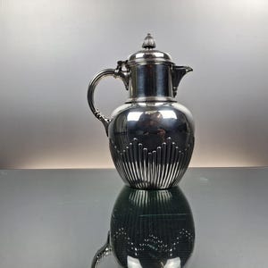 Puede incluir: Una jarra de color plateado con asa, boquilla y tapa. La jarra tiene un cuerpo bulboso con líneas verticales y un patrón decorativo. La jarra es muy reflectante, con su reflejo visible debajo.