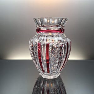 May include: A clear and ruby red cut glass vase featuring a flared rim, a horizontal band near the top, and vertical panels alternating with diamond-patterned sections. The body of the vase has a faceted surface that reflects light throughout.