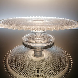 May include: A clear glass cake stand with a scalloped edge and a pedestal base. The stand is reflecting on a dark surface.