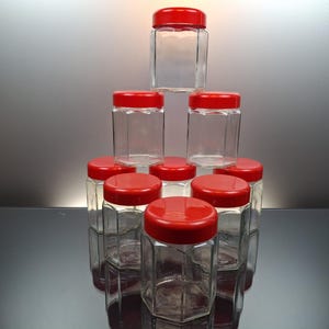 Pode incluir: Uma coleção de nove potes de vidro transparente com corpos hexagonais e tampas de rosca vermelho-brilhante, dispostos em uma pilha piramidal. Cada pote apresenta uma construção de vidro transparente e uma tampa vermelha correspondente.