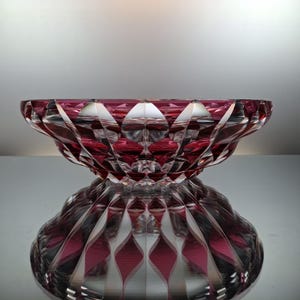 Può includere: Una ciotola decorativa in vetro color rubino con un motivo a taglio diamante. La ciotola si riflette sulla superficie sottostante, creando un'immagine simmetrica. Il design della ciotola presenta linee e sfaccettature che si intersecano.