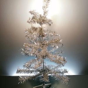 Op de afbeelding: Een zilveren kerstboom van lametta met een stervormige voet. De boom heeft een klassieke conische vorm met meerdere takken. Het lametta reflecteert het licht en creëert een glinsterend effect. Een feestelijke decoratie voor de feestdagen.