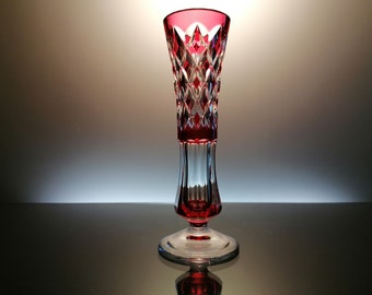May include: A red crystal vase with a diamond pattern and a clear glass base. The vase is on a reflective surface.