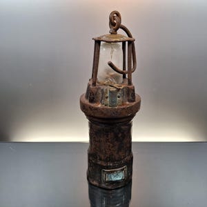 Op de afbeelding: Een antieke, roestige metalen mijnwerkerslamp met een heldere glazen behuizing. De lamp heeft een handvat en een lus aan de bovenkant. De basis is cilindrisch en heeft een kleine rechthoekige plaat. De lamp is een vintage item.