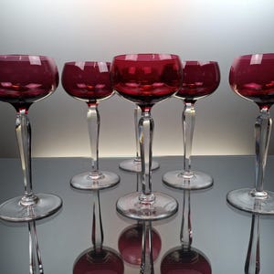 Può includere: Sei bicchieri da vino a stelo trasparenti con una tonalità rosso scuro. I bicchieri sono disposti a semicerchio su una superficie riflettente.