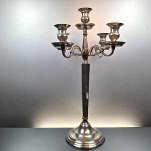 Peut inclure: Un chandelier argenté à cinq bougeoirs. Le chandelier a une tige longue et élancée et une base ronde. Les bougeoirs sont disposés en gradins, avec un au centre et quatre autour.