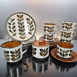 May include: A vintage ceramic dish set featuring a white background with brown and green leaf designs. The set includes plates, cups, saucers, a creamer, and a sugar bowl. The rims and interiors have a brown trim.
