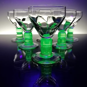 Op de afbeelding: Set van zes heldere glazen drinkglazen met groene voetjes. De glazen hebben een uniek, geometrisch ontwerp en zijn in een rij op een zwart oppervlak gerangschikt.