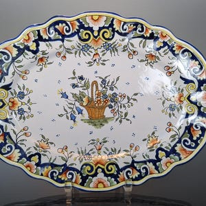 Pode incluir: Travessa oval de cerâmica com fundo branco e desenhos florais azuis, amarelos e laranja. O centro da travessa apresenta uma cesta de flores.