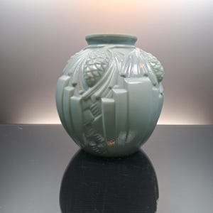 Puede incluir: Un jarrón esférico de vidrio color verde salvia claro con un patrón en relieve de piñas y agujas de pino. El jarrón tiene un acabado liso y brillante con una abertura circular ancha en la parte superior y un cuerpo redondeado hacia la base.