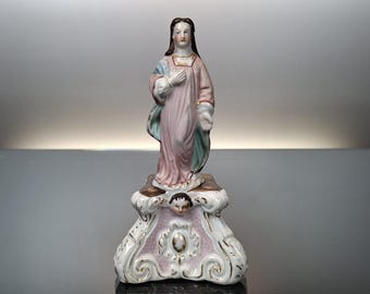 Puede incluir: Una figurita de porcelana de la Virgen María de pie sobre una base decorativa. La figurita está pintada en tonos de rosa, azul y blanco. La base está decorada con detalles dorados.