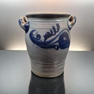 Op de afbeelding: Een keramische vaas met een lichtgrijze basis en een iets donkerdere rand. De vaas heeft twee handvatten en is versierd met blauwe abstracte ontwerpen. De vaas heeft een cilindrische vorm en wordt waarschijnlijk gebruikt voor decoratieve doeleinden.