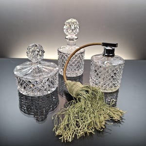 Puede incluir: Tres frascos de perfume de cristal transparente y un tarro pequeño con tapa. Los frascos tienen patrones de corte de diamante y tapones decorativos. También hay un atomizador de perfume vintage con una borla verde.