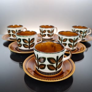Pode incluir: Um conjunto de seis chávenas e pires de café vintage. As chávenas têm um exterior branco com um padrão de folhas castanhas e um interior castanho. Os pires e as bordas das chávenas são castanhos. O conjunto está disposto sobre uma superfície refletora.