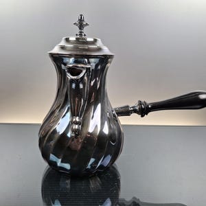 Op de afbeelding: Een zilverkleurige koffiepot met een zwarte handgreep en deksel. De pot heeft een spiraalvormig ontwerp op de body en een kleine tuit. De handgreep is aan de zijkant bevestigd.