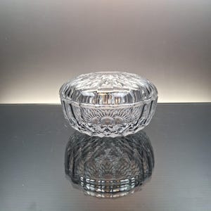 Op de afbeelding: Een ronde, doorzichtige glazen sieradendoos met een afneembaar deksel. De buitenkant is voorzien van een gefacetteerd, geometrisch diamantgeslepen patroon dat het licht opvangt, wat zorgt voor een textuur op zowel de basis als het bolle deksel.