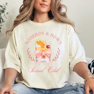 May include: A woman wearing a light beige t-shirt with the text "Bourbon & Bows Social Club" and a pink bow graphic. The graphic features a glass of bourbon with a cherry and orange slice.