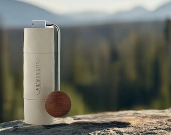 May include: A cylindrical off-white manual coffee grinder with a metallic silver handle and a round dark brown wooden knob. The body features the embossed text COMANDANTE with wing icons on the side, and a smooth, matte finish throughout the exterior.