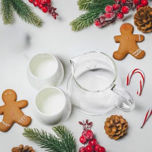 Puede incluir: Una cafetera de vidrio con asa y dos tazas de cerámica blanca. La cafetera y las tazas están llenas de un líquido blanco. Hay galletas de jengibre, bastones de caramelo y ramas de pino con bayas rojas sobre una superficie blanca.