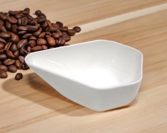 May include: A white ceramic scoop with a rounded, triangular shape. The scoop is empty and sitting on a wooden surface. A pile of coffee beans is in the background.