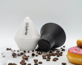 Könnte beinhalten: Ein weißer Keramik-Pour-Over-Kaffeefilter mit dem Aufdruck "LilyDrip" neben einem schwarzen Plastik-Pour-Over-Kaffeefilter. Beide Filter liegen auf einer weißen Oberfläche mit verstreuten Kaffeebohnen.