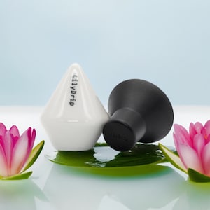 K&ouml;nnte beinhalten: Zwei Keramik-Flaschenverschl&uuml;sse, einer wei&szlig; und einer schwarz, mit dem Text "LilyDrip" auf dem wei&szlig;en Verschluss. Die Verschl&uuml;sse liegen auf einem gr&uuml;nen Blatt mit rosa Bl&uuml;ten auf beiden Seiten.