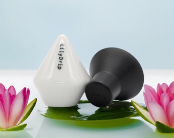 May include: Two ceramic bottle stoppers, one white and one black, with the text "LilyDrip" on the white stopper. The stoppers are sitting on a green leaf with pink flowers on either side.
