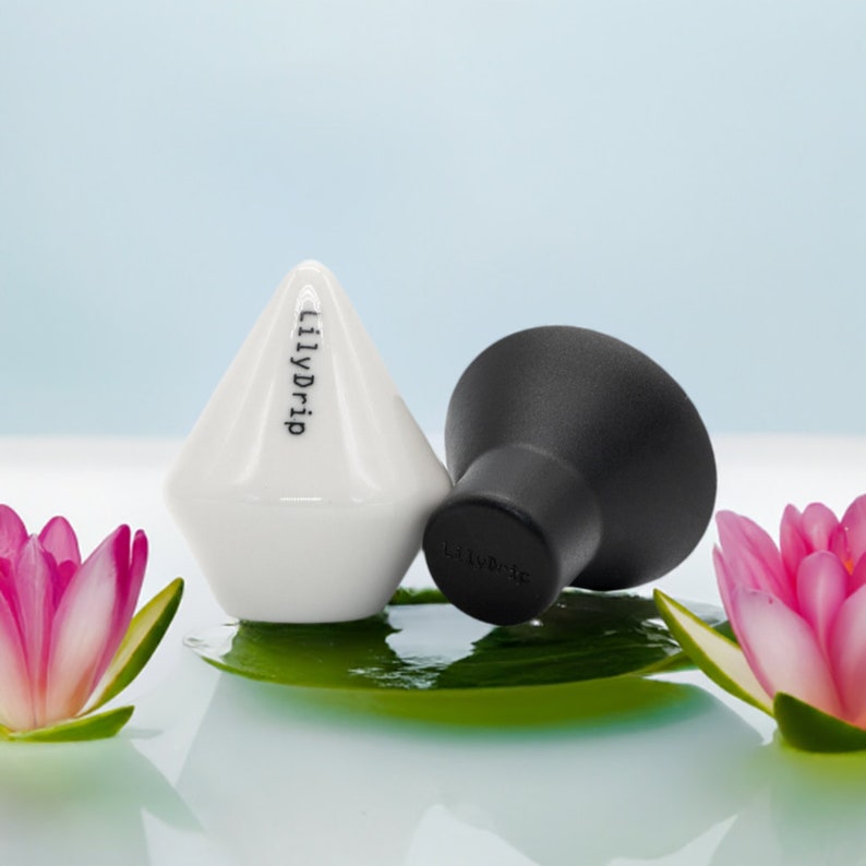 K&ouml;nnte beinhalten: Zwei Keramik-Flaschenverschl&uuml;sse, einer wei&szlig; und einer schwarz, mit dem Text "LilyDrip" auf dem wei&szlig;en Verschluss. Die Verschl&uuml;sse liegen auf einem gr&uuml;nen Blatt mit rosa Bl&uuml;ten auf beiden Seiten.