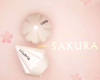 Könnte beinhalten: Zwei weiße Keramikbehälter mit dem Text "Lilly Drip" darauf. Die Behälter sind in Form einer Blume und eines Kegels geformt. Die Behälter befinden sich auf einem rosa Hintergrund mit einem Kirschblütenmuster.