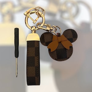 May include: A brown and black checkered keychain with a gold metal ring and a brown leather bow. The keychain is shaped like a cartoon mouse head.
