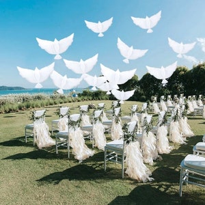 May include: An outdoor wedding ceremony setup with white chairs adorned with fabric and greenery. White dove decorations float in the sky above the chairs, creating a peaceful and elegant atmosphere. The scene is set on a green lawn with a view of the ocean.