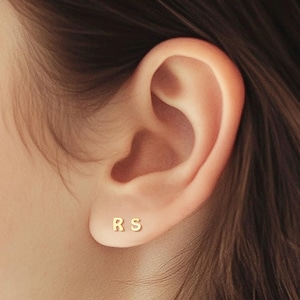 Può includere: Un paio di orecchini iniziali in oro con le lettere "RS" su di essi. Gli orecchini sono indossati nell'orecchio sinistro di una persona con i capelli castani.