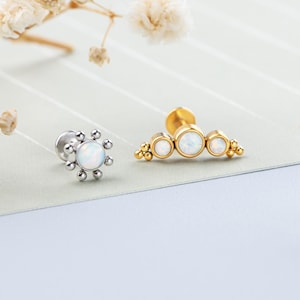 Op de afbeelding: Er worden twee sieraden getoond. Eén is een zilveren oorbel met een bloemachtig ontwerp en een witte opaal in het midden. De andere is een gouden stuk met drie opaalstenen. De achtergrond is een lichtblauwe en groene ondergrond met witte bloemen.