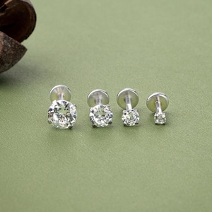 May include: Four silver-colored stud earrings with clear, round, sparkling gemstones of varying sizes. The earrings are arranged in a row against a muted green background, showcasing their brilliance. The studs are designed for ear or lip piercings.