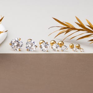 Può includere: Una collezione di orecchini a lobo dorati con pietre di zirconia cubica trasparente. Gli orecchini variano in dimensioni, con i più grandi a sinistra e che diminuiscono verso destra. Presentati su una superficie bianca e marrone.