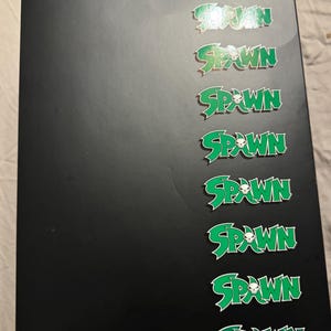 Könnte beinhalten: Eine schwarze Box mit mehreren grünen "Spawn"-Logos. Jedes Logo enthält eine Totenkopf-Grafik. Die Logos sind in einem sich wiederholenden Muster an der Seite angeordnet. Die Logos sind weiß umrandet.