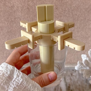 Puede incluir: Un molde de plástico beige para paletas con un tallo central y múltiples brazos que se extienden hacia afuera, diseñado para sostener palitos de paleta. El molde se coloca dentro de un vaso transparente. El diseño permite hacer múltiples paletas a la vez.