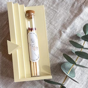 Könnte beinhalten: Ein Glasröhrchen gefüllt mit Streichhölzern, mit einem Korken und einem Etikett mit floraler Linienkunst und dem Text "Thank You So Much". Das Röhrchen liegt in einem hellgelben Halter mit einem dreieckigen Akzent.