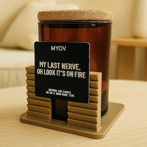 Può includere: Una candela di vetro marrone con coperchio in sughero, appoggiata su un supporto beige. Un cartello nero davanti recita "MY LAST NERVE, OH LOOK IT'S ON FIRE" con il marchio "MYOV". La candela è una candela naturale.