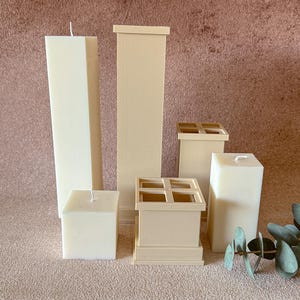 Peut inclure: Un assortiment de bougies ivoire de différentes formes et tailles. La collection comprend des bougies hautes, rectangulaires et carrées, certaines avec un motif en forme de croix sur le dessus. Les bougies sont disposées sur une surface texturée brun clair.