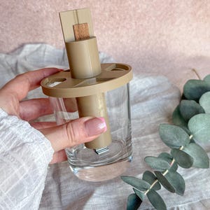 Puede incluir: Un portavelas de vidrio transparente con un inserto de plástico color beige y una mecha de madera. El soporte está parcialmente lleno de líquido. El diseño incluye una base circular y una parte superior rectangular. El fondo es rosa y gris claro.