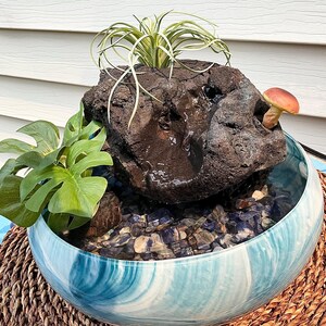 May include: A decorative indoor water fountain featuring a dark, textured rock formation with a small mushroom and greenery. The fountain sits in a blue and white swirled ceramic bowl filled with pebbles. The bowl rests on a woven mat.