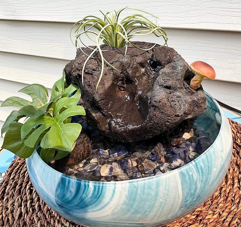 May include: A decorative indoor water fountain featuring a dark, textured rock formation with a small mushroom and greenery. The fountain sits in a blue and white swirled ceramic bowl filled with pebbles. The bowl rests on a woven mat.