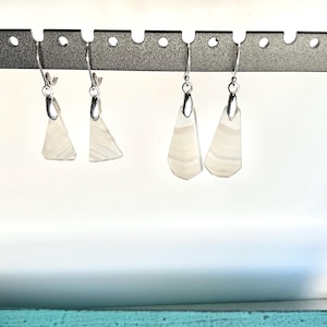 May include: Four pairs of dangle earrings displayed on a silver metal earring holder. The earrings feature a silver hook and a white, geometric, triangular pendant with subtle, horizontal, light grey stripes. The earrings are displayed against a white background.